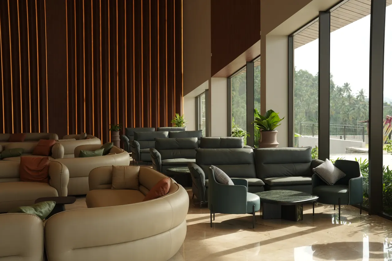 Guest lounge with curved cream sofas and teal armchairs beside floor-to-ceiling windows overlooking palm trees